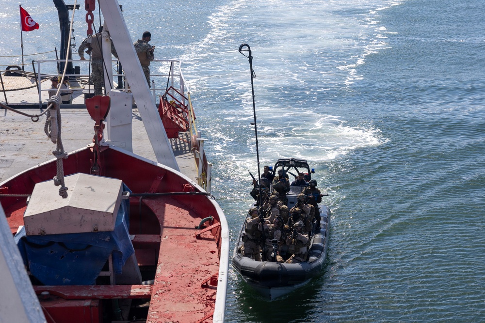 A military or law enforcement tactical operation is underway involving a rigid inflatable boat (RIB) carrying multiple armed personnel approaching a larger vessel. The larger ship displays a Tunisian flag, suggesting this may be a maritime interdiction or boarding exercise in Tunisian waters. Personnel on both the RIB and the ship's deck are equipped with tactical gear and weapons.