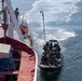 A military or law enforcement tactical operation is underway involving a rigid inflatable boat (RIB) carrying multiple armed personnel approaching a larger vessel. The larger ship displays a Tunisian flag, suggesting this may be a maritime interdiction or boarding exercise in Tunisian waters. Personnel on both the RIB and the ship's deck are equipped with tactical gear and weapons.
