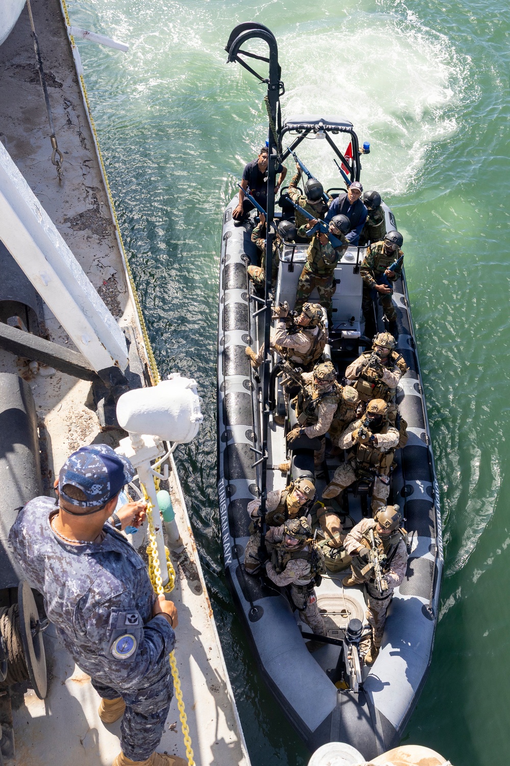 An overhead view shows a rigid-hull inflatable boat (RHIB) carrying multiple armed military personnel in full tactical gear approaching the side of a larger vessel. A sailor in blue camouflage uniform stands on the deck of the larger ship, managing a chain or line. The scene appears to depict a maritime boarding or visit, board, search, and seizure (VBSS) training exercise or operation.