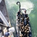 An overhead view shows a rigid-hull inflatable boat (RHIB) carrying multiple armed military personnel in full tactical gear approaching the side of a larger vessel. A sailor in blue camouflage uniform stands on the deck of the larger ship, managing a chain or line. The scene appears to depict a maritime boarding or visit, board, search, and seizure (VBSS) training exercise or operation.