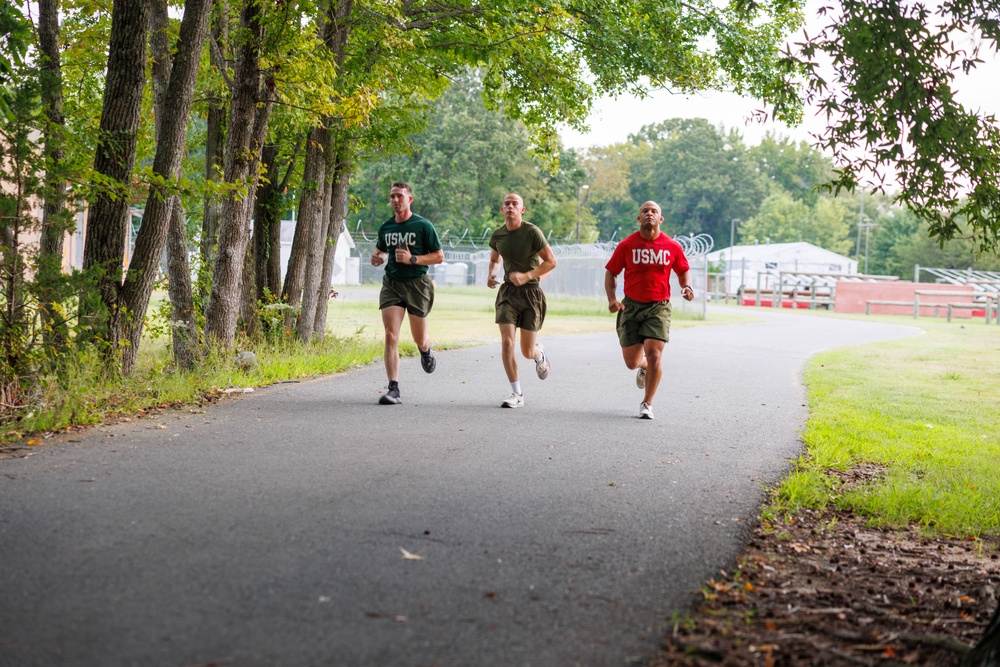 Three individuals wearing USMC (United States Marine Corps) athletic gear are running together on a paved path, likely on a military base. Two are wearing green USMC shirts and one is wearing a red USMC shirt, all paired with olive/khaki shorts. The setting appears to be a well-maintained base facility with trees lining the path and military structures visible in the background.