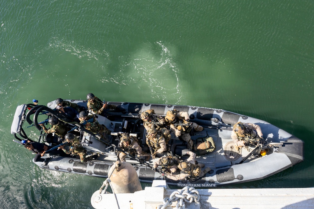 An aerial/overhead view of a gray rigid inflatable boat (RIB) marked 'TUNISIAN NAVY' carrying multiple armed military personnel in tactical gear. The personnel appear to be conducting a maritime boarding or insertion exercise, with soldiers in mixed camouflage uniforms and helmets. The boat is positioned alongside what appears to be a larger vessel, visible at the bottom of the frame.