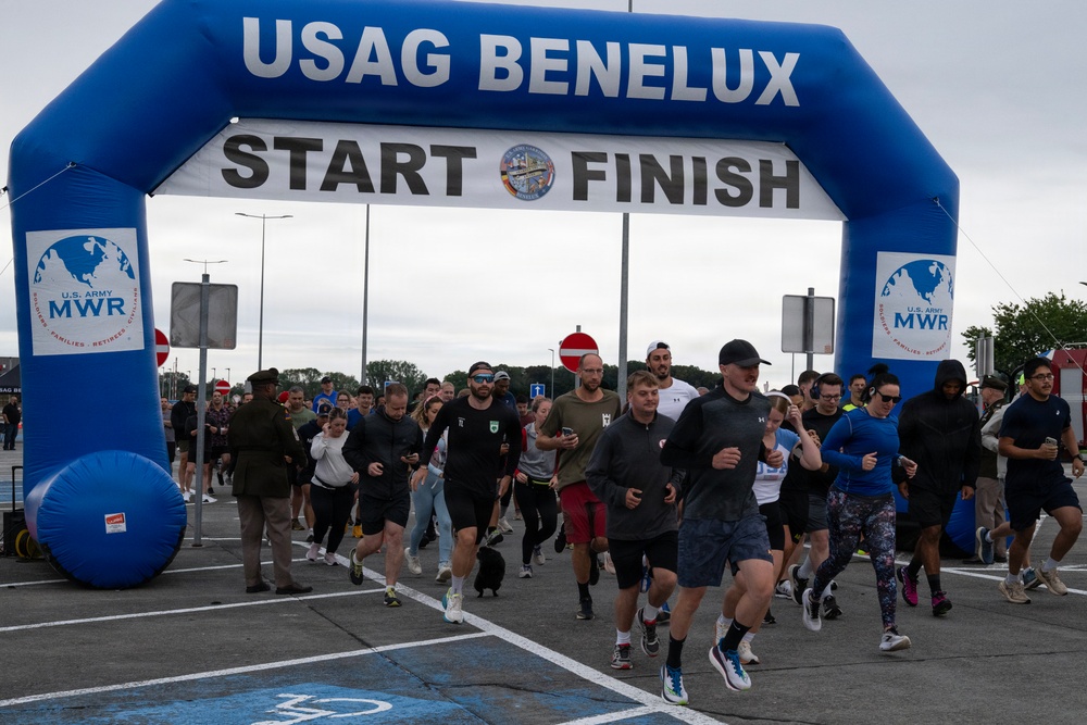 A large group of participants is crossing the start/finish line of a running event organized by USAG Benelux (U.S. Army Garrison Benelux). The event is sponsored by U.S. Army MWR (Morale, Welfare, and Recreation) as indicated by signage on the inflatable arch. Participants of various ages and fitness attire are actively running through the arch on what appears to be an overcast day.