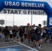 A large group of participants is crossing the start/finish line of a running event organized by USAG Benelux (U.S. Army Garrison Benelux). The event is sponsored by U.S. Army MWR (Morale, Welfare, and Recreation) as indicated by signage on the inflatable arch. Participants of various ages and fitness attire are actively running through the arch on what appears to be an overcast day.