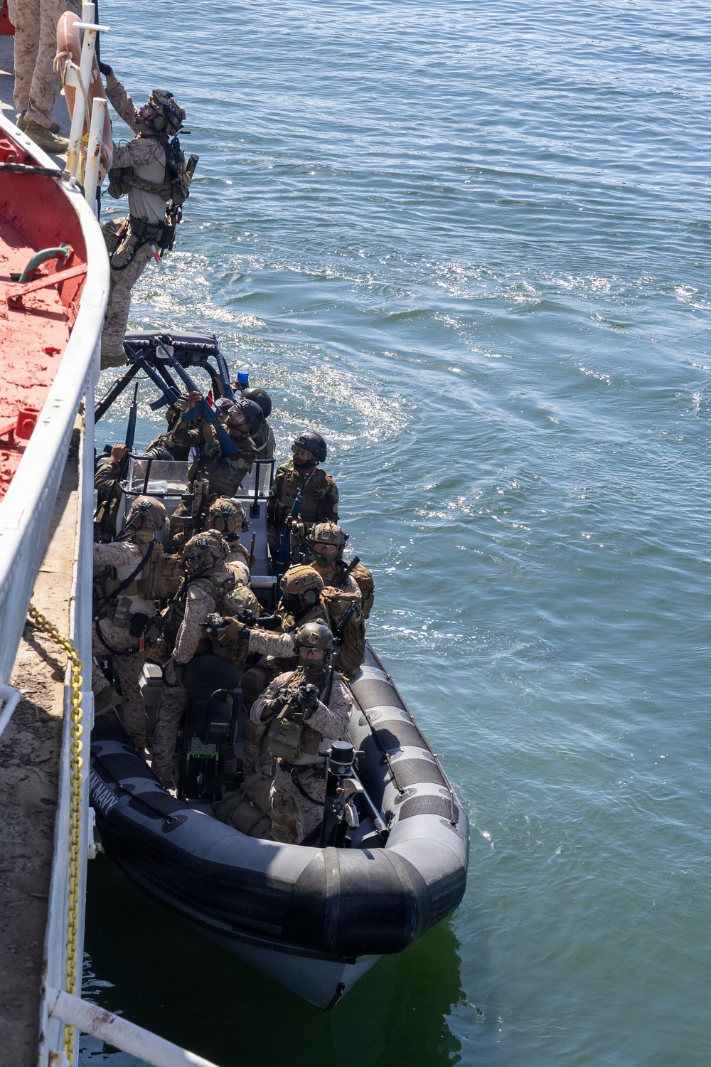 Military personnel in full tactical gear are conducting a maritime boarding operation from a rigid-hull inflatable boat (RHIB) alongside a larger vessel. One operator is climbing up the side of the ship while others in the RHIB maintain security positions with weapons. This appears to be a visit, board, search, and seizure (VBSS) training exercise or operation.
