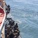 Military personnel in full tactical gear are conducting a maritime boarding operation from a rigid-hull inflatable boat (RHIB) alongside a larger vessel. One operator is climbing up the side of the ship while others in the RHIB maintain security positions with weapons. This appears to be a visit, board, search, and seizure (VBSS) training exercise or operation.