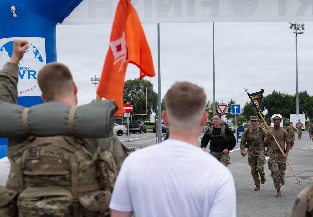 Military personnel are approaching a finish line during what appears to be a ruck march or military fitness event. Soldiers in camouflage uniforms are running toward the finish area, with one carrying a unit guidon flag. Spectators and other soldiers are cheering near a blue inflatable arch bearing 'Army' and 'VR' branding associated with Army Veteran/Retiree services.