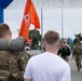Military personnel are approaching a finish line during what appears to be a ruck march or military fitness event. Soldiers in camouflage uniforms are running toward the finish area, with one carrying a unit guidon flag. Spectators and other soldiers are cheering near a blue inflatable arch bearing 'Army' and 'VR' branding associated with Army Veteran/Retiree services.