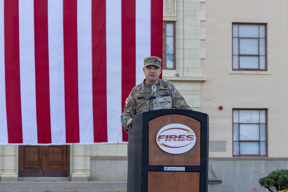 A U.S. Army officer in OCP (Operational Camouflage Pattern) uniform is speaking at a podium labeled 'FIRES' during an outdoor military ceremony. A large American flag is displayed as a backdrop against what appears to be a military installation building. The officer appears to hold at least a one-star general rank based on visible insignia.