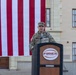 A U.S. Army officer in OCP (Operational Camouflage Pattern) uniform is speaking at a podium labeled 'FIRES' during an outdoor military ceremony. A large American flag is displayed as a backdrop against what appears to be a military installation building. The officer appears to hold at least a one-star general rank based on visible insignia.