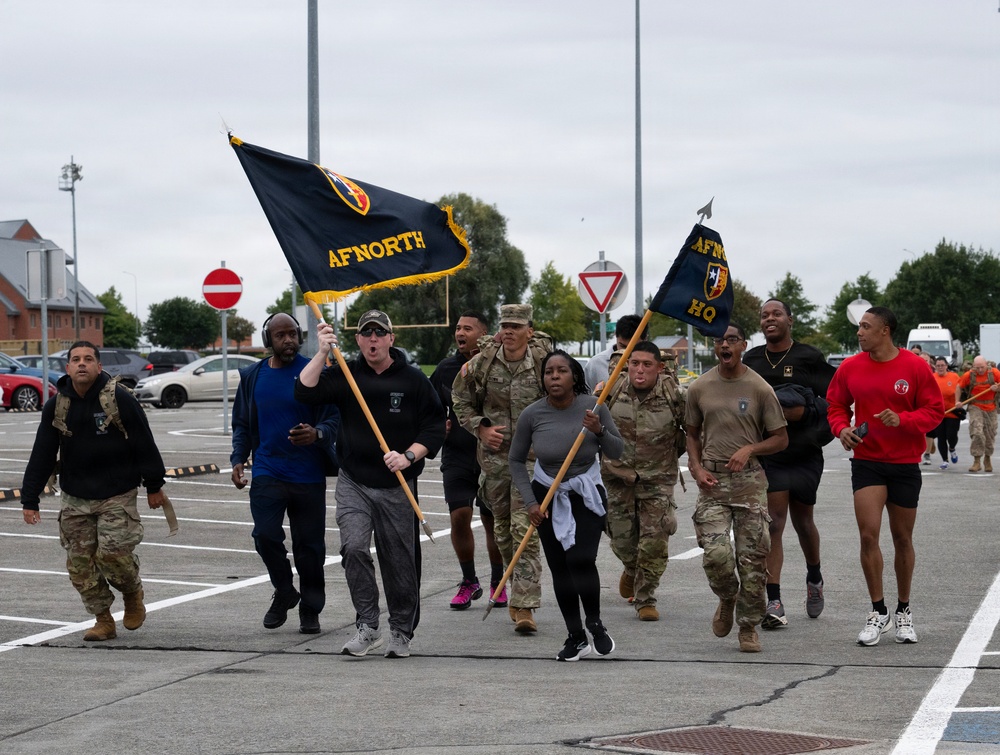 A group of military personnel and civilians are running or marching together in a parking lot while carrying unit guidon flags marked 'AFNORTH' and 'AFNORTH HQ'. The participants are dressed in a mix of military camouflage uniforms and civilian athletic wear, suggesting a unit physical fitness event or run. The setting appears to be a military installation, likely in Europe based on the visible European-style road signs.