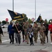 A group of military personnel and civilians are running or marching together in a parking lot while carrying unit guidon flags marked 'AFNORTH' and 'AFNORTH HQ'. The participants are dressed in a mix of military camouflage uniforms and civilian athletic wear, suggesting a unit physical fitness event or run. The setting appears to be a military installation, likely in Europe based on the visible European-style road signs.