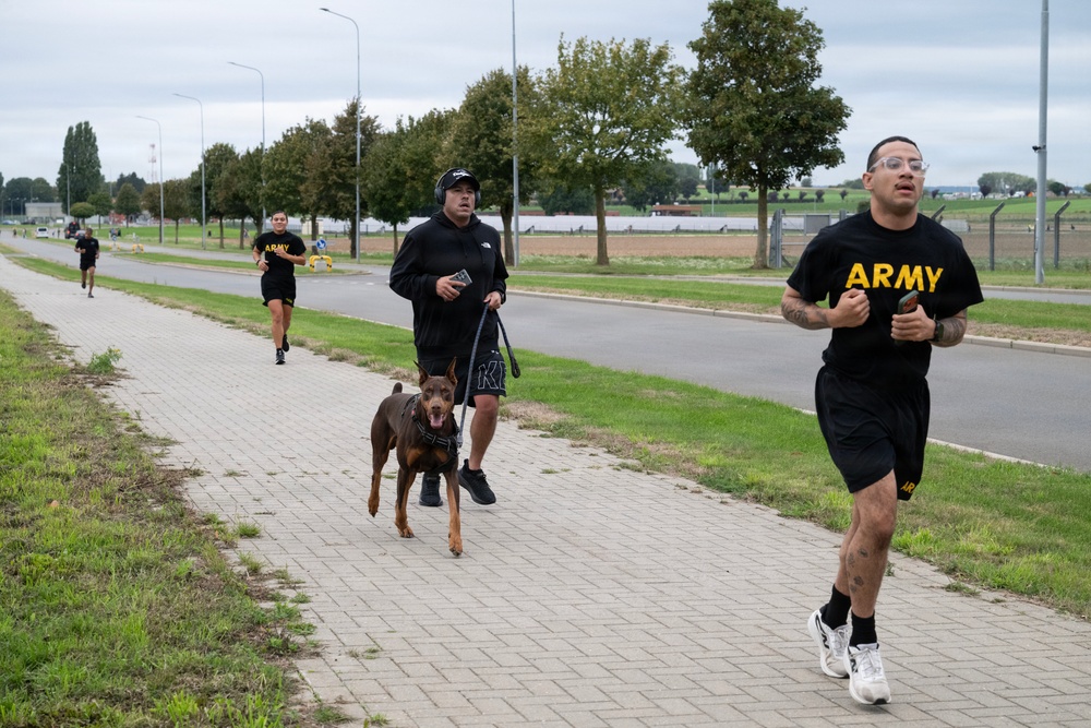 A group of U.S. Army personnel are jogging along a paved sidewalk path, likely on or near a military installation. One individual in the middle of the group is running with a brown Doberman Pinscher on a leash. The setting appears to be a well-maintained base or park road lined with trees and street lamps under an overcast sky.