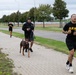 A group of U.S. Army personnel are jogging along a paved sidewalk path, likely on or near a military installation. One individual in the middle of the group is running with a brown Doberman Pinscher on a leash. The setting appears to be a well-maintained base or park road lined with trees and street lamps under an overcast sky.