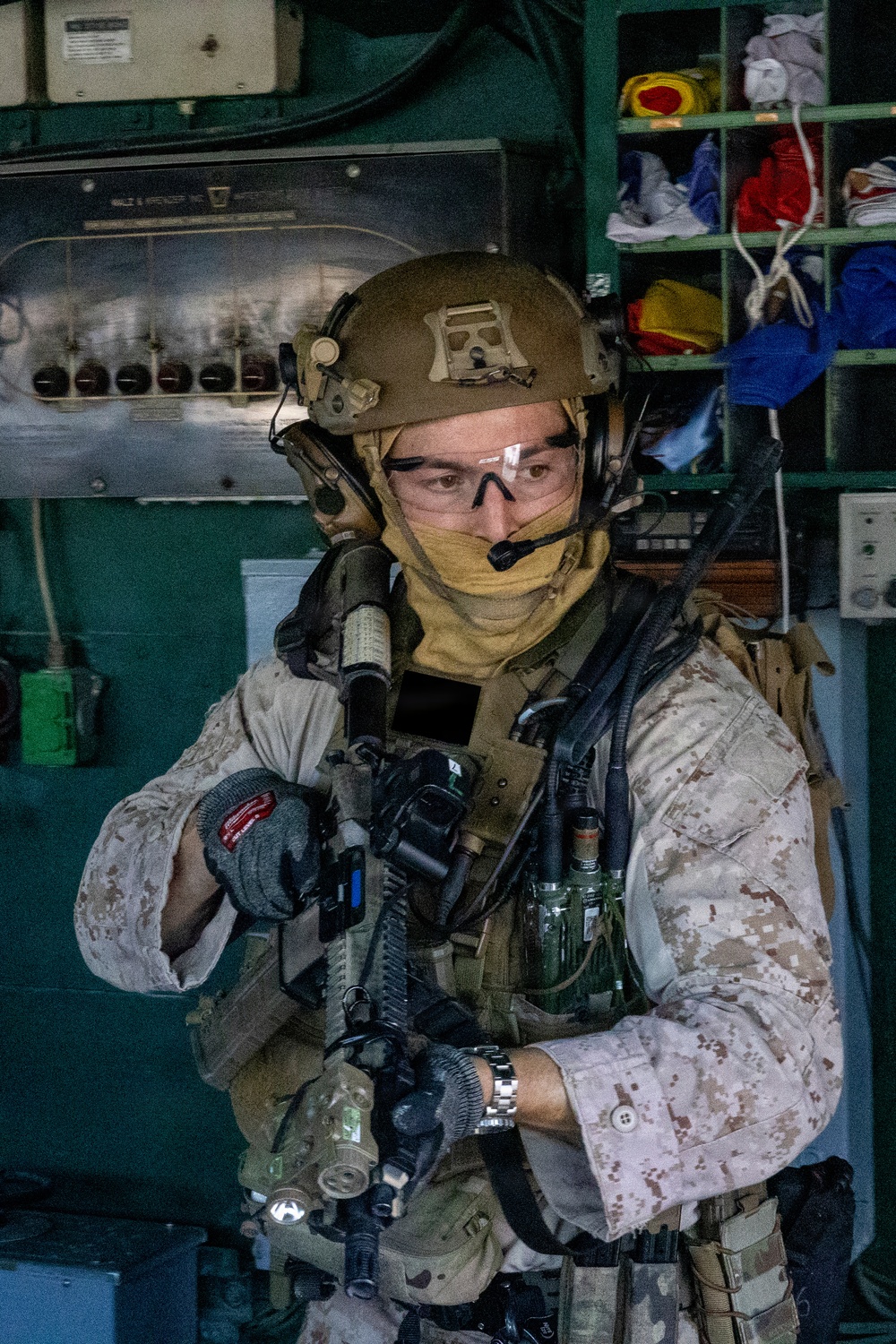 A heavily equipped military operator is photographed inside what appears to be a ship's interior compartment or engine room. The individual is wearing full tactical gear including a ballistic helmet, communications headset, protective eyewear, face covering, body armor, and camouflage uniform. The operator is holding a weapon system with multiple attachments and appears to be in a ready or clearing posture.