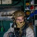 A heavily equipped military operator is photographed inside what appears to be a ship's interior compartment or engine room. The individual is wearing full tactical gear including a ballistic helmet, communications headset, protective eyewear, face covering, body armor, and camouflage uniform. The operator is holding a weapon system with multiple attachments and appears to be in a ready or clearing posture.