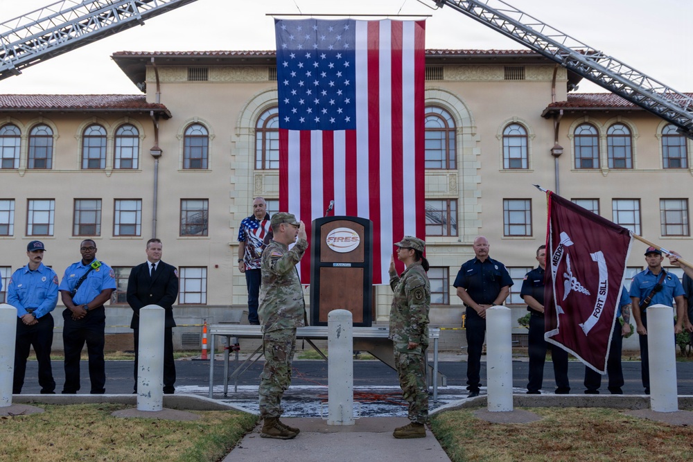 A military ceremony is taking place in front of a large building at Fort Sill, with two soldiers in camouflage uniforms exchanging salutes in front of a podium bearing the 'FIRES' insignia. A large American flag is suspended between two fire truck ladder extensions in the background. Firefighters and other personnel in blue uniforms stand in formation as witnesses to the event.