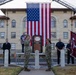 A military ceremony is taking place in front of a large building at Fort Sill, with two soldiers in camouflage uniforms exchanging salutes in front of a podium bearing the 'FIRES' insignia. A large American flag is suspended between two fire truck ladder extensions in the background. Firefighters and other personnel in blue uniforms stand in formation as witnesses to the event.