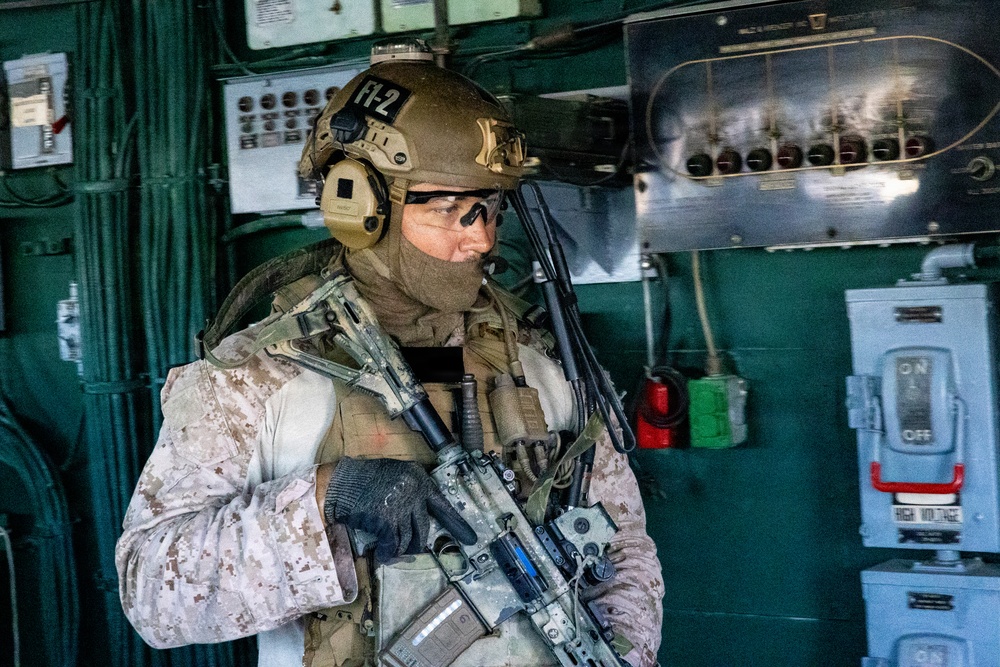 A heavily equipped military operator is positioned inside what appears to be a ship's engine room or control space. The individual is wearing MARPAT desert camouflage uniform with full tactical gear including a tan helmet marked 'FT-2', communications equipment, and is holding an assault rifle. The background shows green-painted industrial panels with switches, gauges, and safety equipment consistent with a naval vessel interior.