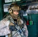 A heavily equipped military operator is positioned inside what appears to be a ship's engine room or control space. The individual is wearing MARPAT desert camouflage uniform with full tactical gear including a tan helmet marked 'FT-2', communications equipment, and is holding an assault rifle. The background shows green-painted industrial panels with switches, gauges, and safety equipment consistent with a naval vessel interior.