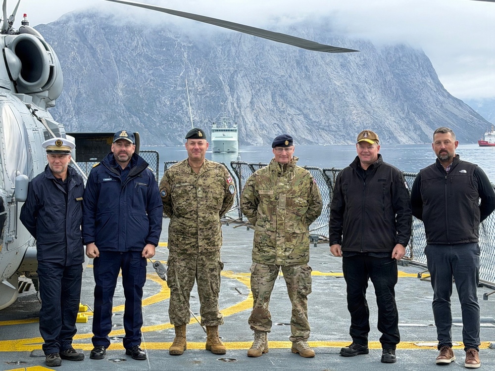 Six military and civilian personnel are posed for a group photograph on the flight deck of a naval vessel, with a helicopter visible to the left. The setting appears to be a Nordic fjord environment, with dramatic steep rocky cliffs and misty mountains in the background. At least two vessels are visible in the fjord behind the group.