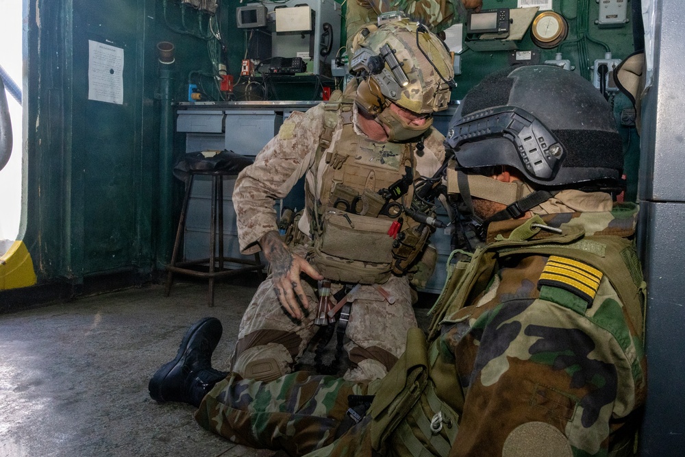 Two military personnel are inside what appears to be a ship or vessel interior. One soldier in MARPAT desert camouflage and full tactical gear is kneeling and appears to be administering first aid or conducting a tactical assessment on a second individual wearing woodland camouflage with gold rank insignia on the shoulder. The setting suggests a training exercise or operational scenario aboard a naval vessel.