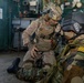 Two military personnel are inside what appears to be a ship or vessel interior. One soldier in MARPAT desert camouflage and full tactical gear is kneeling and appears to be administering first aid or conducting a tactical assessment on a second individual wearing woodland camouflage with gold rank insignia on the shoulder. The setting suggests a training exercise or operational scenario aboard a naval vessel.