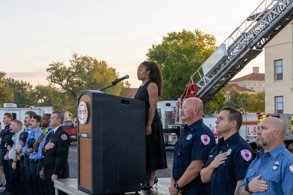 A formal outdoor ceremony is taking place, likely a memorial or commemorative event, where a woman in a black dress is singing or speaking at a podium while firefighters and officials stand at attention with hands over their hearts. A fire department ladder truck is visible in the background with its aerial ladder extended. The podium displays what appears to be a fire department emblem or logo.