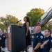 A formal outdoor ceremony is taking place, likely a memorial or commemorative event, where a woman in a black dress is singing or speaking at a podium while firefighters and officials stand at attention with hands over their hearts. A fire department ladder truck is visible in the background with its aerial ladder extended. The podium displays what appears to be a fire department emblem or logo.