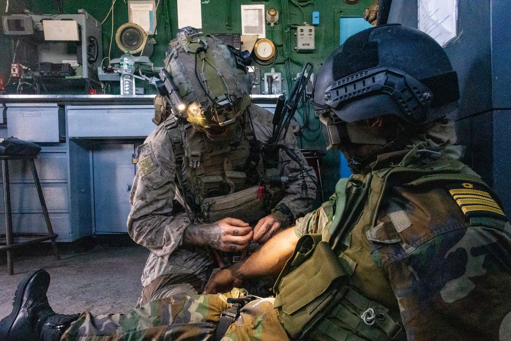 Two military personnel are shown in what appears to be an indoor tactical environment, possibly a ship or facility control room. One soldier in desert/woodland camouflage with a helmet-mounted light is administering what appears to be tactical combat casualty care (TCCC) or first aid to a second individual wearing a black ballistic helmet and camouflage uniform with rank insignia. The scene suggests a training exercise or real-world medical assistance scenario.