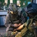 Two military personnel are shown in what appears to be an indoor tactical environment, possibly a ship or facility control room. One soldier in desert/woodland camouflage with a helmet-mounted light is administering what appears to be tactical combat casualty care (TCCC) or first aid to a second individual wearing a black ballistic helmet and camouflage uniform with rank insignia. The scene suggests a training exercise or real-world medical assistance scenario.