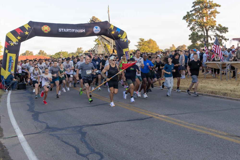 A large group of runners is crossing the start/finish line of an organized road race, likely held on a military installation based on visible insignia and signage. The inflatable arch prominently displays 'START/FINISH' along with what appears to be Fort Hall or similar military base branding and MWR (Morale, Welfare, and Recreation) logos. A lead runner carries a red flag/pennant while participants of various ages and fitness levels surge forward at the race start.
