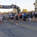 A large group of runners is crossing the start/finish line of an organized road race, likely held on a military installation based on visible insignia and signage. The inflatable arch prominently displays 'START/FINISH' along with what appears to be Fort Hall or similar military base branding and MWR (Morale, Welfare, and Recreation) logos. A lead runner carries a red flag/pennant while participants of various ages and fitness levels surge forward at the race start.