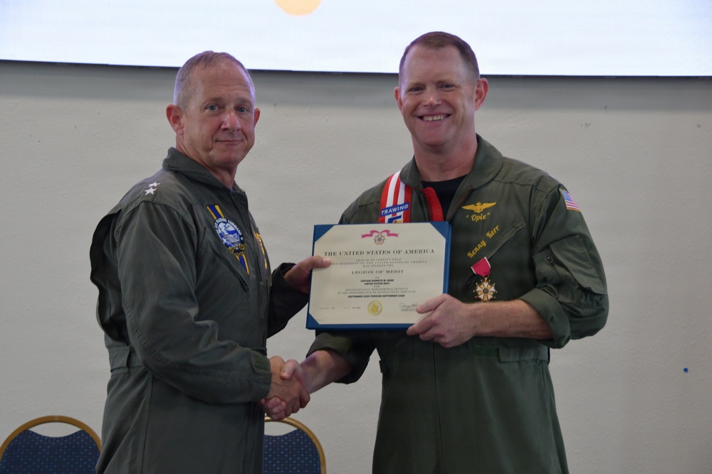 Two U.S. Navy officers in olive green flight suits are shaking hands during a formal military award ceremony. The officer on the right is receiving a Legion of Merit certificate and medal, with a red-and-white ribbon/lanyard around his neck. The ceremony appears to be taking place indoors, likely in a conference or meeting room.