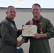 Two U.S. Navy officers in olive green flight suits are shaking hands during a formal military award ceremony. The officer on the right is receiving a Legion of Merit certificate and medal, with a red-and-white ribbon/lanyard around his neck. The ceremony appears to be taking place indoors, likely in a conference or meeting room.