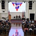 A formal military ceremony is taking place in a large hall, featuring the TRAWING 5 (Training Air Wing Five) insignia displayed on a large projection screen. An officer is speaking at a podium on stage while two uniformed personnel are seated behind him, with an American flag and what appears to be a Marine Corps flag flanking the stage. A large audience of military personnel and civilians fills the seating area.