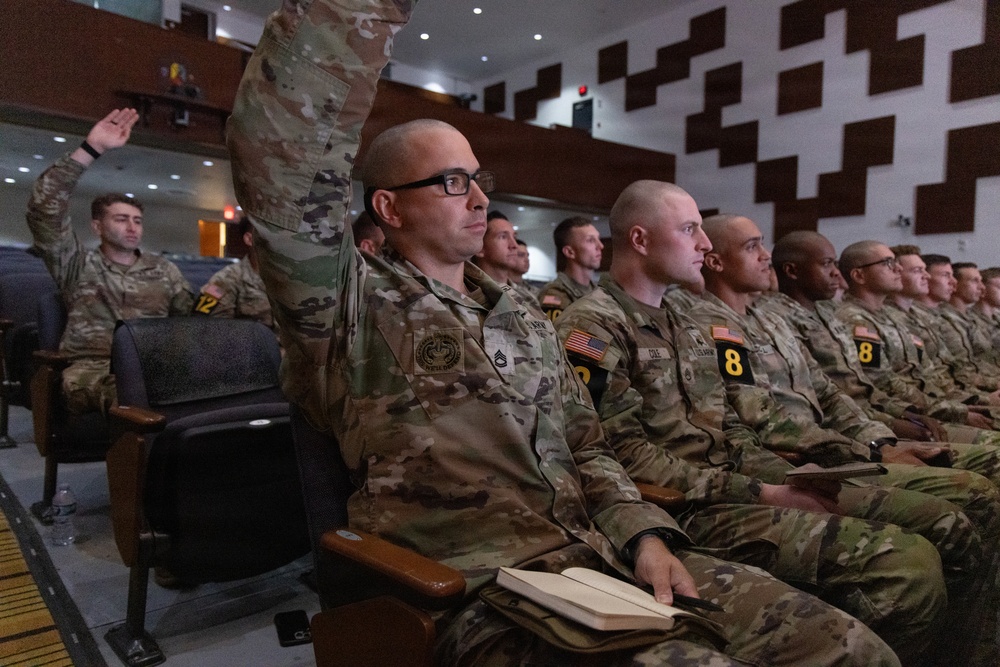 A group of U.S. Army soldiers in OCP (Operational Camouflage Pattern) uniforms are seated in an auditorium-style briefing room. One soldier in the foreground is raising his hand, likely asking a question or responding during a briefing or training session. Several soldiers are wearing numbered placards, suggesting a competition, evaluation, or organized training event.
