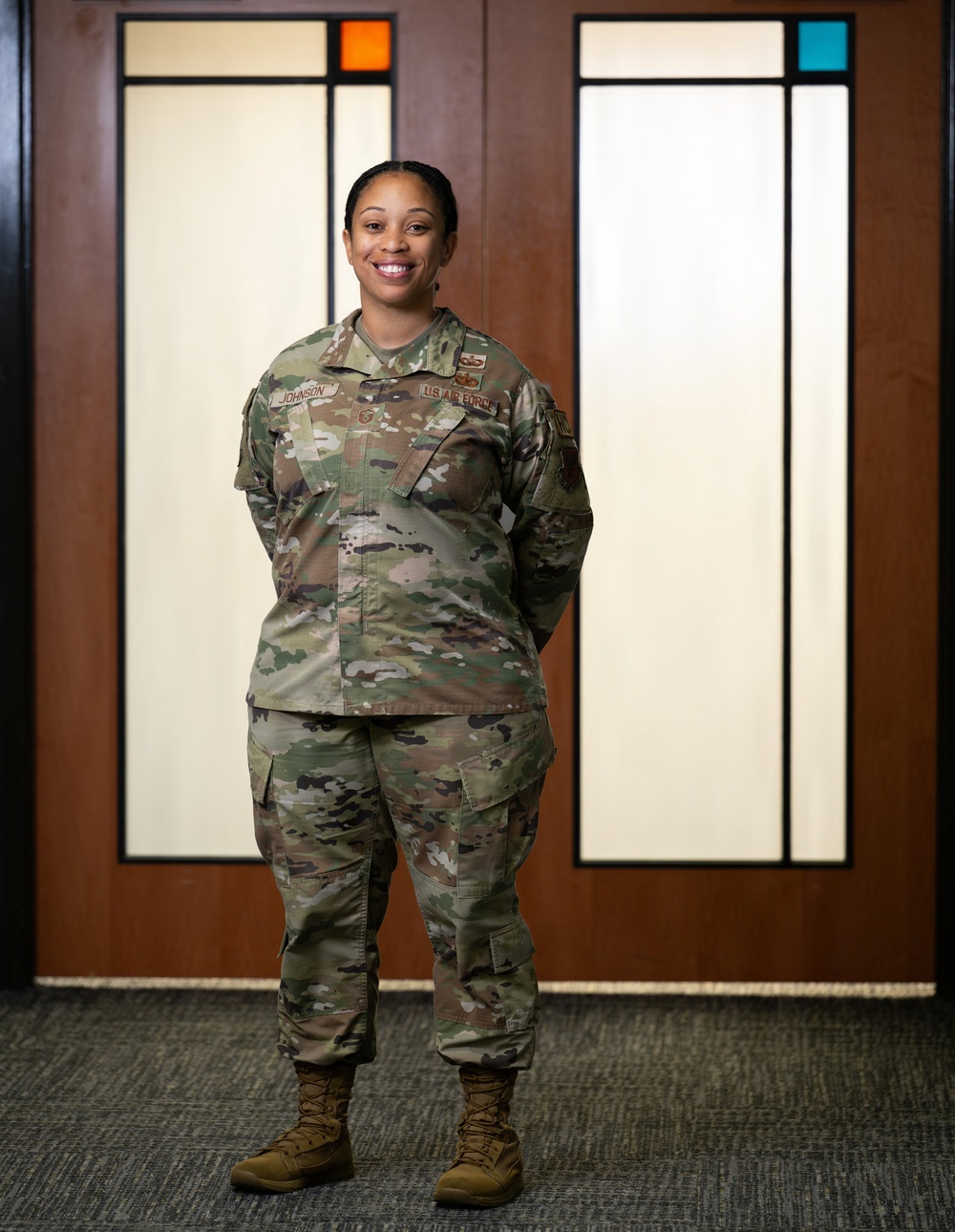 A U.S. Air Force service member named Johnson stands in a full-body portrait pose, smiling, wearing the Operational Camouflage Pattern (OCP) uniform with visible name tape and branch tape. The individual is photographed indoors in front of decorative wooden panel doors featuring colored glass accents in orange and teal. The image appears to be an official or semi-official military portrait.