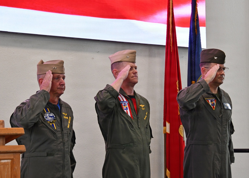 Three military officers in flight suits are rendering a salute during a formal ceremony. They are standing in front of what appears to be a large projected American flag backdrop, with military color guard flags visible to their right. The setting appears to be an indoor military change of command or similar official ceremony.