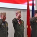 Three military officers in flight suits are rendering a salute during a formal ceremony. They are standing in front of what appears to be a large projected American flag backdrop, with military color guard flags visible to their right. The setting appears to be an indoor military change of command or similar official ceremony.