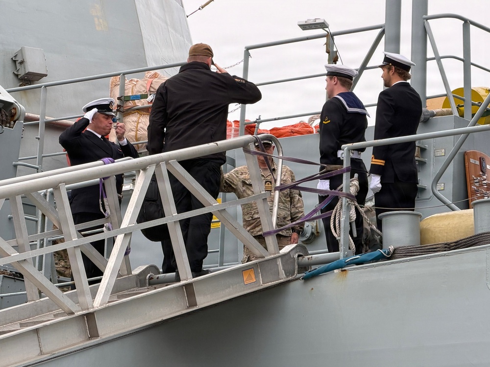 The image shows a formal naval boarding or arrival ceremony on the gangway of a warship. Several personnel in naval dress uniforms are rendering salutes, while others in military camouflage and civilian attire are also present on the gangplank. The scene appears to depict an official visit or dignitary arrival at a naval vessel.