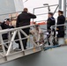 The image shows a formal naval boarding or arrival ceremony on the gangway of a warship. Several personnel in naval dress uniforms are rendering salutes, while others in military camouflage and civilian attire are also present on the gangplank. The scene appears to depict an official visit or dignitary arrival at a naval vessel.