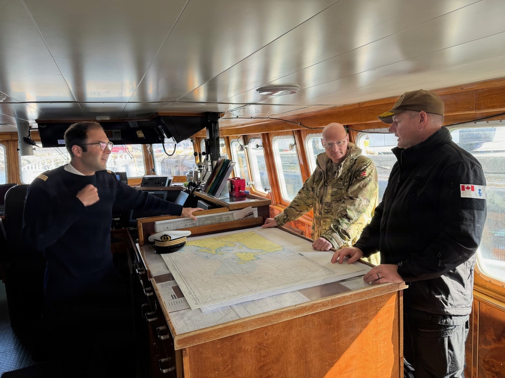Three individuals are gathered on the bridge of a vessel reviewing nautical charts spread across a navigation table. One person wearing a dark naval uniform with shoulder epaulettes appears to be briefing the other two, one of whom wears a multicam/military camouflage uniform with what appears to be a Danish flag patch, and the other wears a black jacket with a Canadian flag patch. The setting is a well-equipped ship's bridge with navigation instruments, windows overlooking a harbor, and wooden cabinetry.