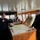 Three individuals are gathered on the bridge of a vessel reviewing nautical charts spread across a navigation table. One person wearing a dark naval uniform with shoulder epaulettes appears to be briefing the other two, one of whom wears a multicam/military camouflage uniform with what appears to be a Danish flag patch, and the other wears a black jacket with a Canadian flag patch. The setting is a well-equipped ship's bridge with navigation instruments, windows overlooking a harbor, and wooden cabinetry.