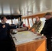 Three individuals are gathered on the bridge of a vessel reviewing nautical charts spread across a navigation table. One person wearing a dark naval uniform with shoulder epaulettes appears to be briefing the other two, one of whom wears a multicam/military camouflage uniform with what appears to be a Danish flag patch, and the other wears a black jacket with a Canadian flag patch. The setting is a well-equipped ship's bridge with navigation instruments, windows overlooking a harbor, and wooden cabinetry.