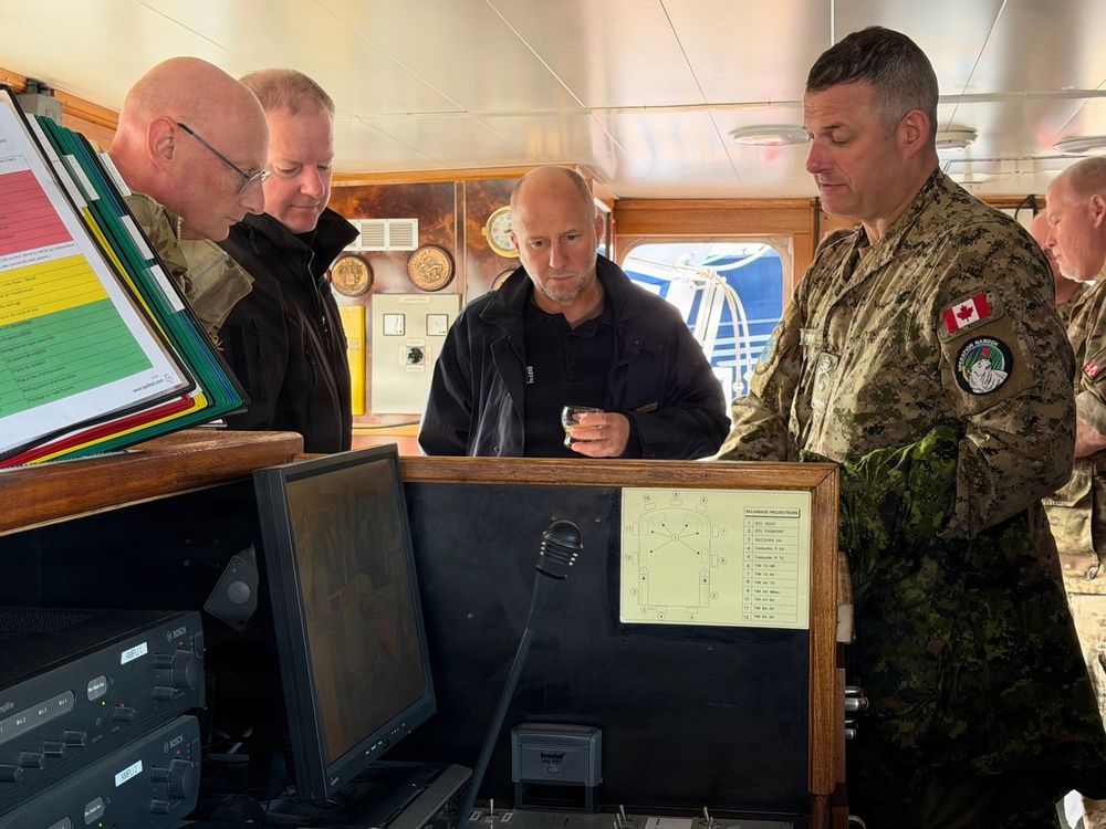 Several military personnel and a civilian are gathered on what appears to be the bridge of a naval or maritime vessel. They are examining navigation equipment and documents at a control console. At least one individual is wearing a Canadian Armed Forces uniform with a visible Canadian flag patch.