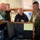 Several military personnel and a civilian are gathered on what appears to be the bridge of a naval or maritime vessel. They are examining navigation equipment and documents at a control console. At least one individual is wearing a Canadian Armed Forces uniform with a visible Canadian flag patch.