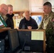 Several military personnel and a civilian are gathered on what appears to be the bridge of a naval or maritime vessel. They are examining navigation equipment and documents at a control console. At least one individual is wearing a Canadian Armed Forces uniform with a visible Canadian flag patch.