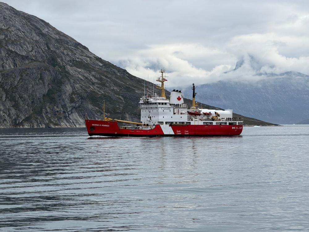 A Canadian Coast Guard vessel named 'CCGS George R. Pearkes' is navigating calm arctic or subarctic waters surrounded by dramatic rocky mountains and overcast skies. The ship displays the distinctive red hull and white superstructure with Canadian maple leaf livery typical of Canadian Coast Guard icebreakers. The scene appears to be in a fjord-like environment, possibly in northern Canada or Greenland.