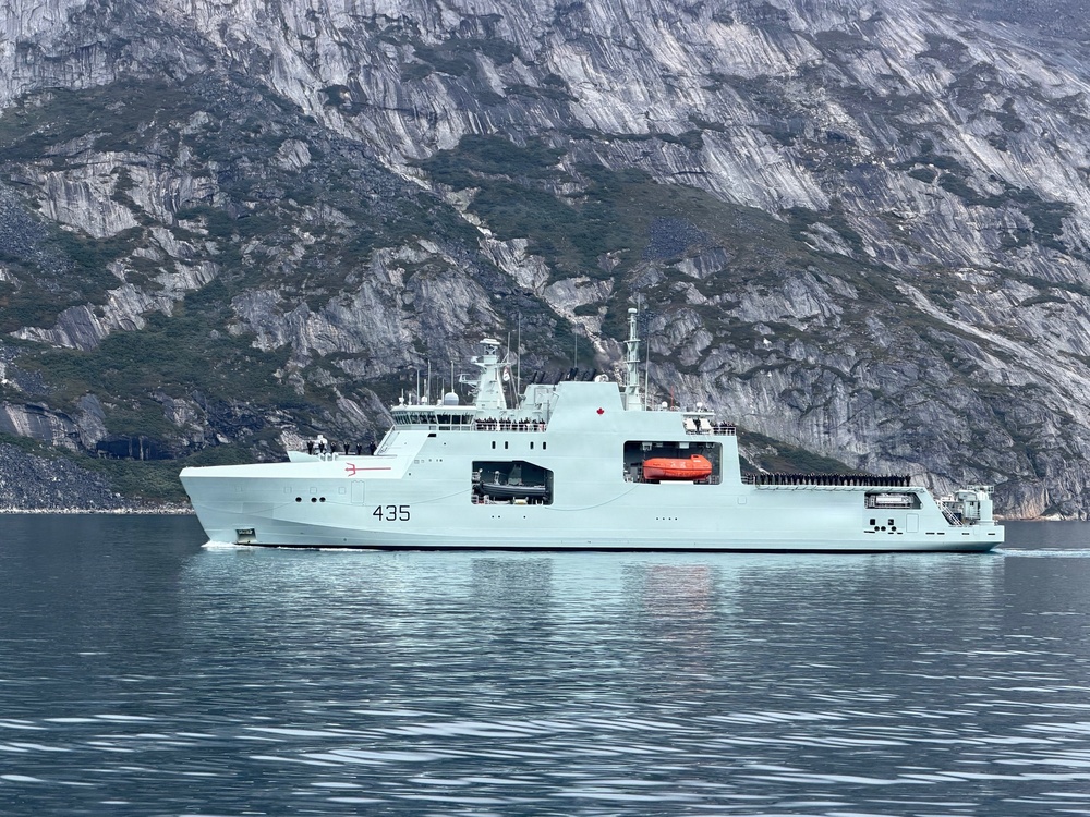 A light grey military vessel bearing hull number 435 and a Canadian maple leaf emblem is underway in calm waters against a dramatic rocky cliff backdrop. The ship appears to be a Canadian Arctic/Offshore Patrol Ship (AOPS), consistent with the Harry DeWolf-class design operated by the Royal Canadian Navy. Personnel are visible lining the upper decks, suggesting a formal or ceremonial transit.
